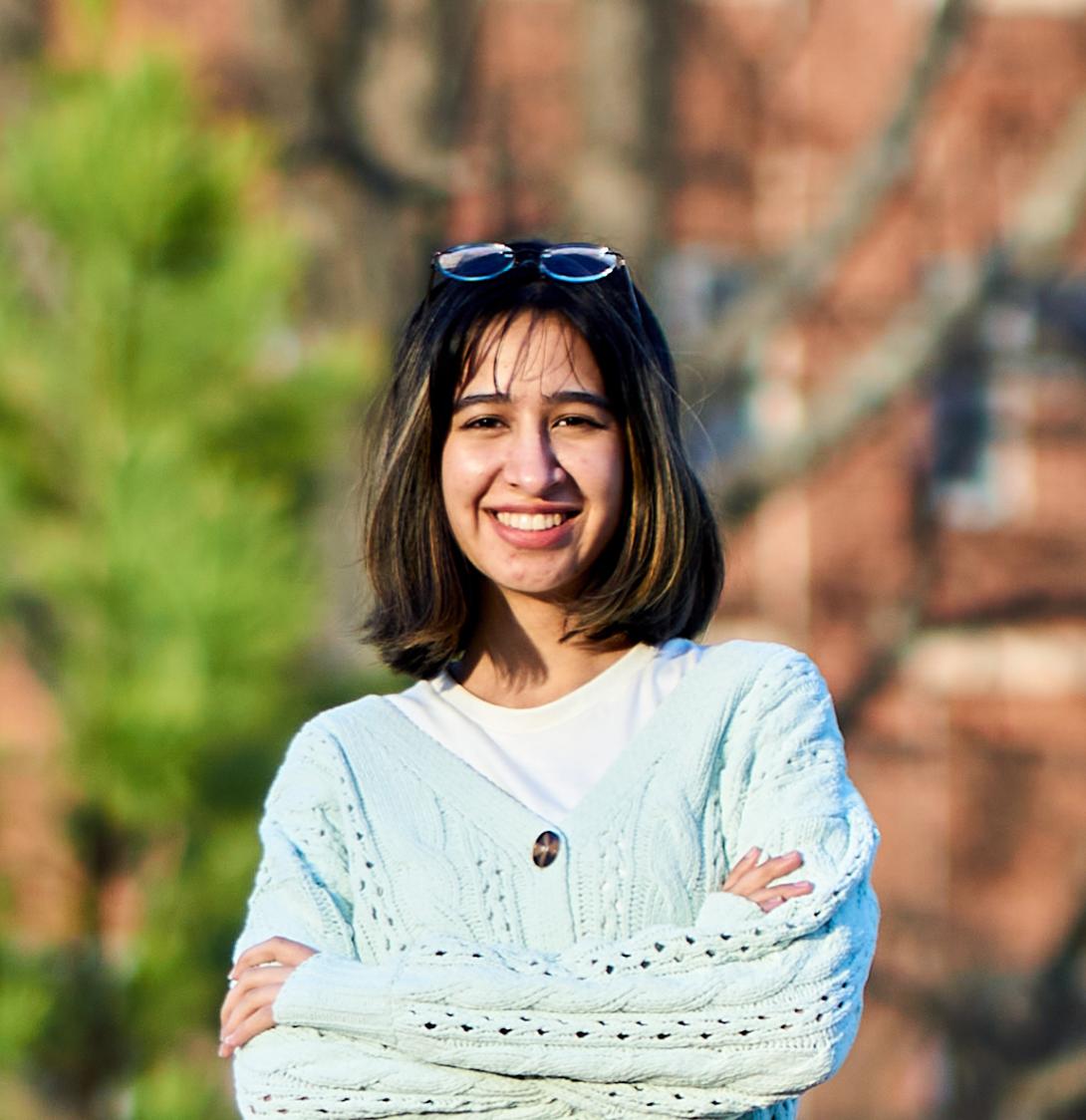 A smiling Azka with shoulder-length dark hair stands outdoors with their arms crossed, wearing a light blue knit cardigan over a white shirt. Spectacles rest on their head. The portrait is taken in warm, late-afternoon sunlight with a softly blurred background of trees and a brick building, creating a natural, welcoming atmosphere.